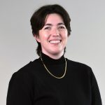 A person with short brown hair, wearing a black textured top and a gold necklace, smiles against a plain light gray background.