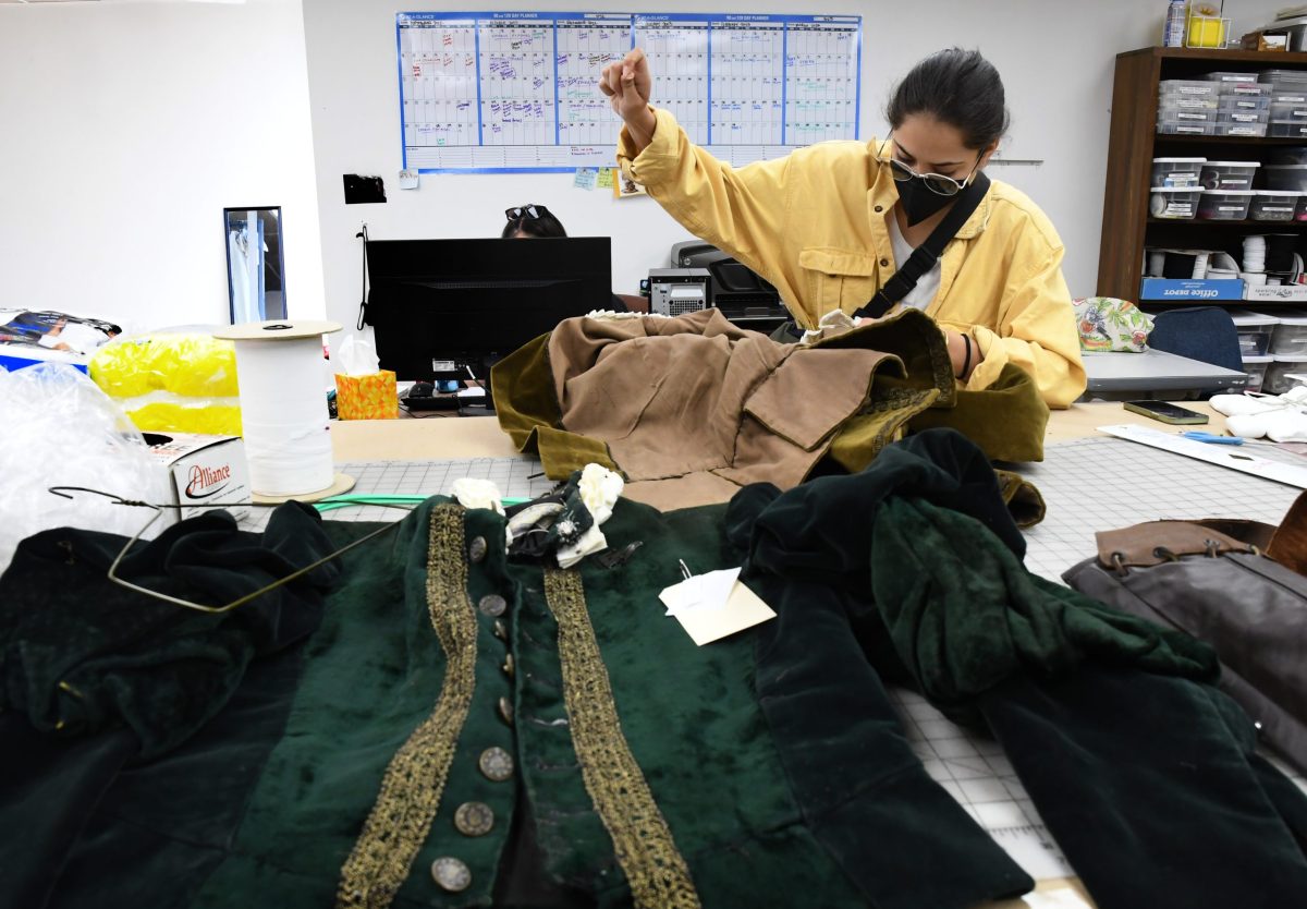 A woman sews a brown coat while an elegant green jacket lays in front ofher.