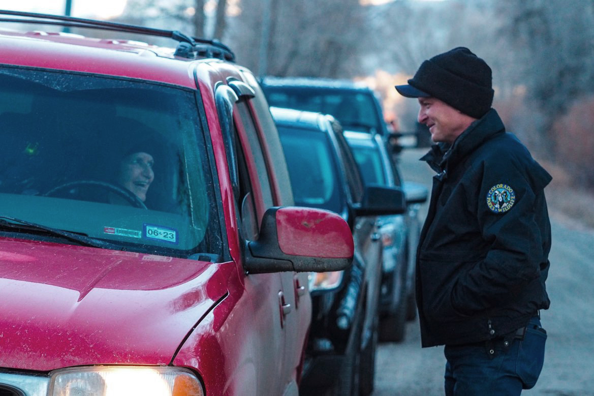 A Colorado Parks and Wildlife worker talks with a woman through the front window of her car as a line of cars wait behind her.