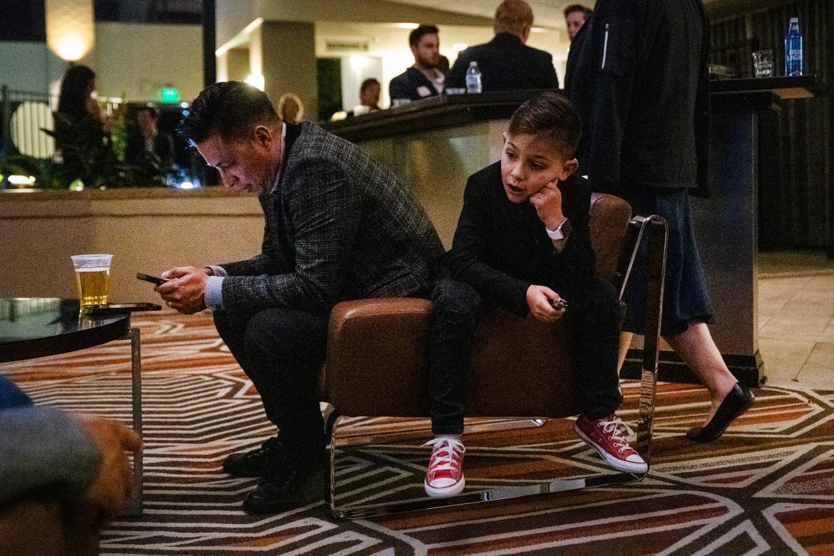 A dad checks his phone while a kid sits on the arm of his chair looking bored.