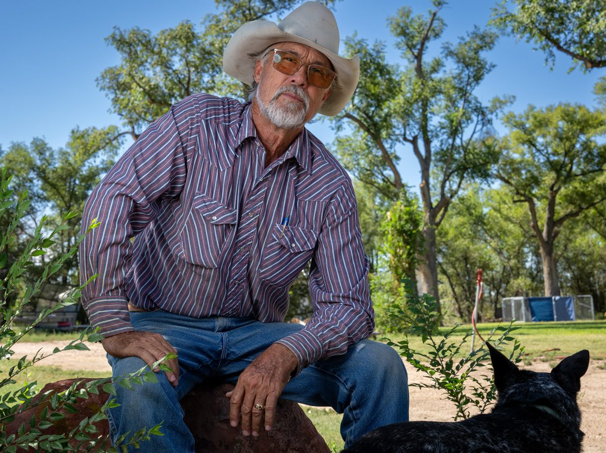 A man wearing a white cowboy hat and a blue striped shirt sits on a rock. His dog is visible at the bottom of the frame