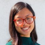 A person with long dark hair and red glasses smiles at the camera, wearing a green sweater in front of a plain light background.
